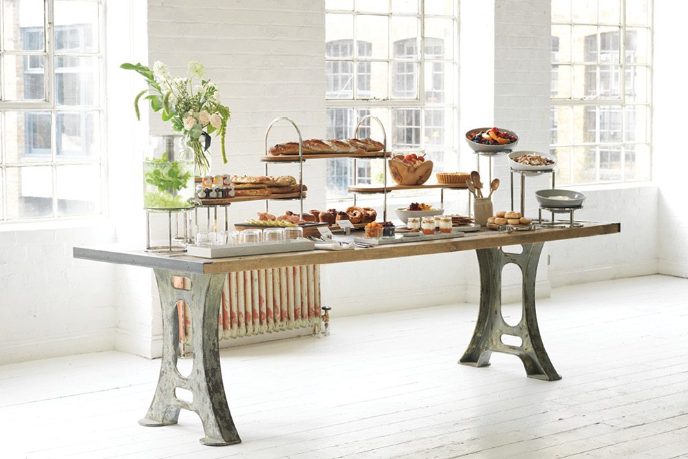 A Craster buffet display: wooden and steel risers with bread, pastries, fruit and yoghurts on a long industrial table in a bright, white room.